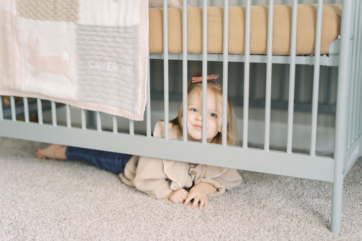 A small child peeks playfully under a white crib in a nursery with gray carpet.