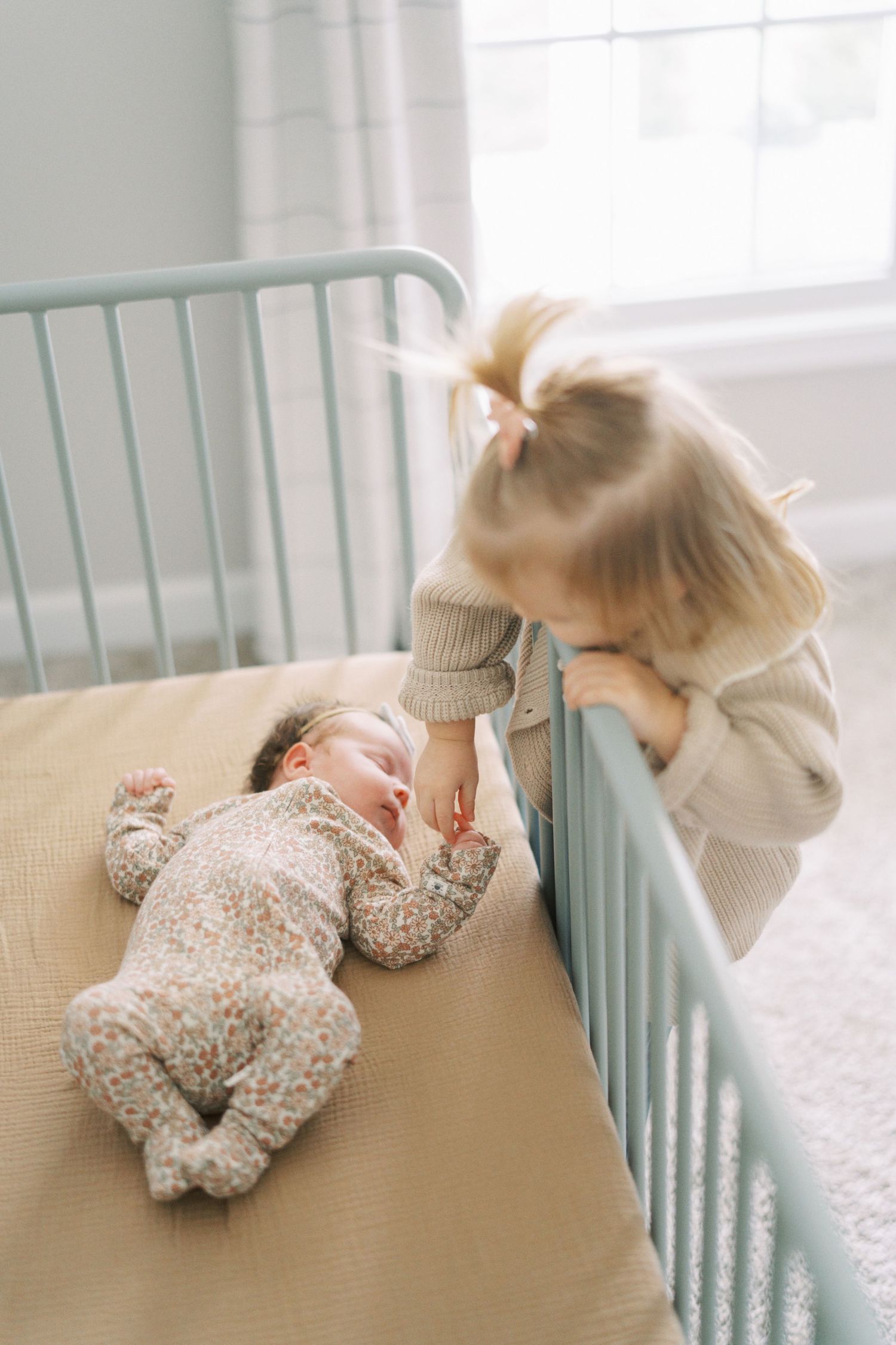 Toddler leaning over crib rail to look at baby lying on beige bedding.
