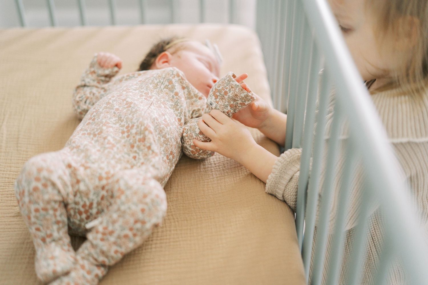 A baby lies on a beige cushion next to crib rails in soft lighting.