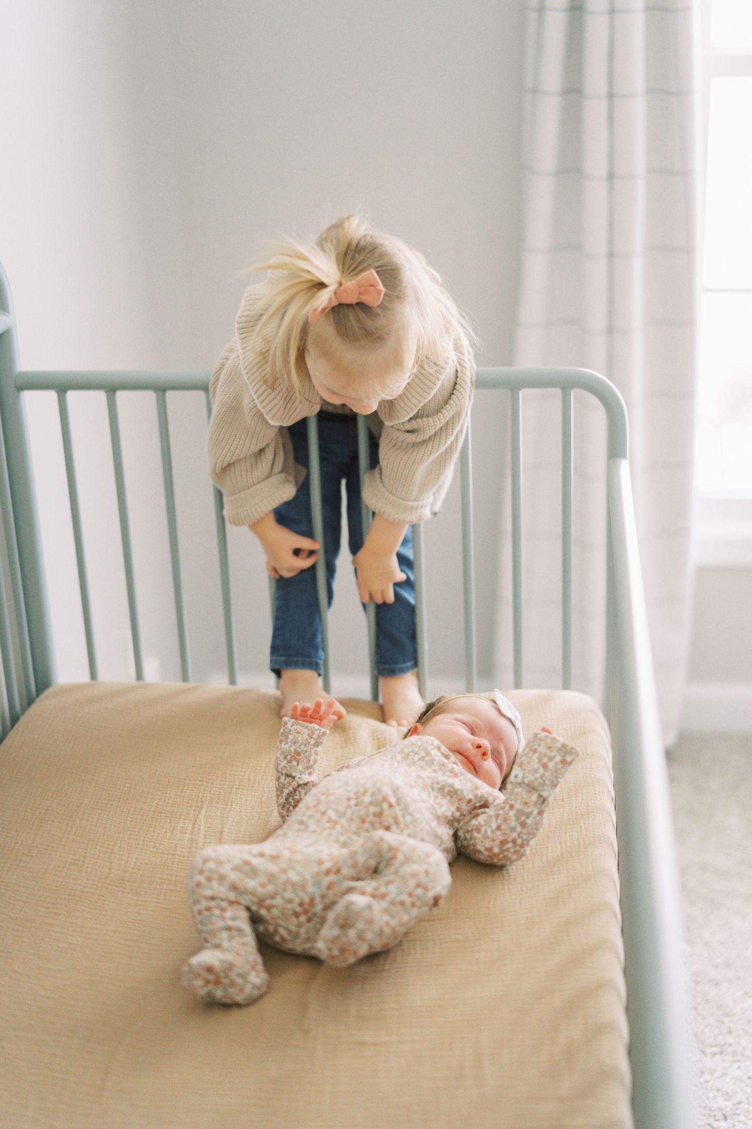 A child in a beige sweater looks down at a baby lying on a bed wearing floral patterned clothing.