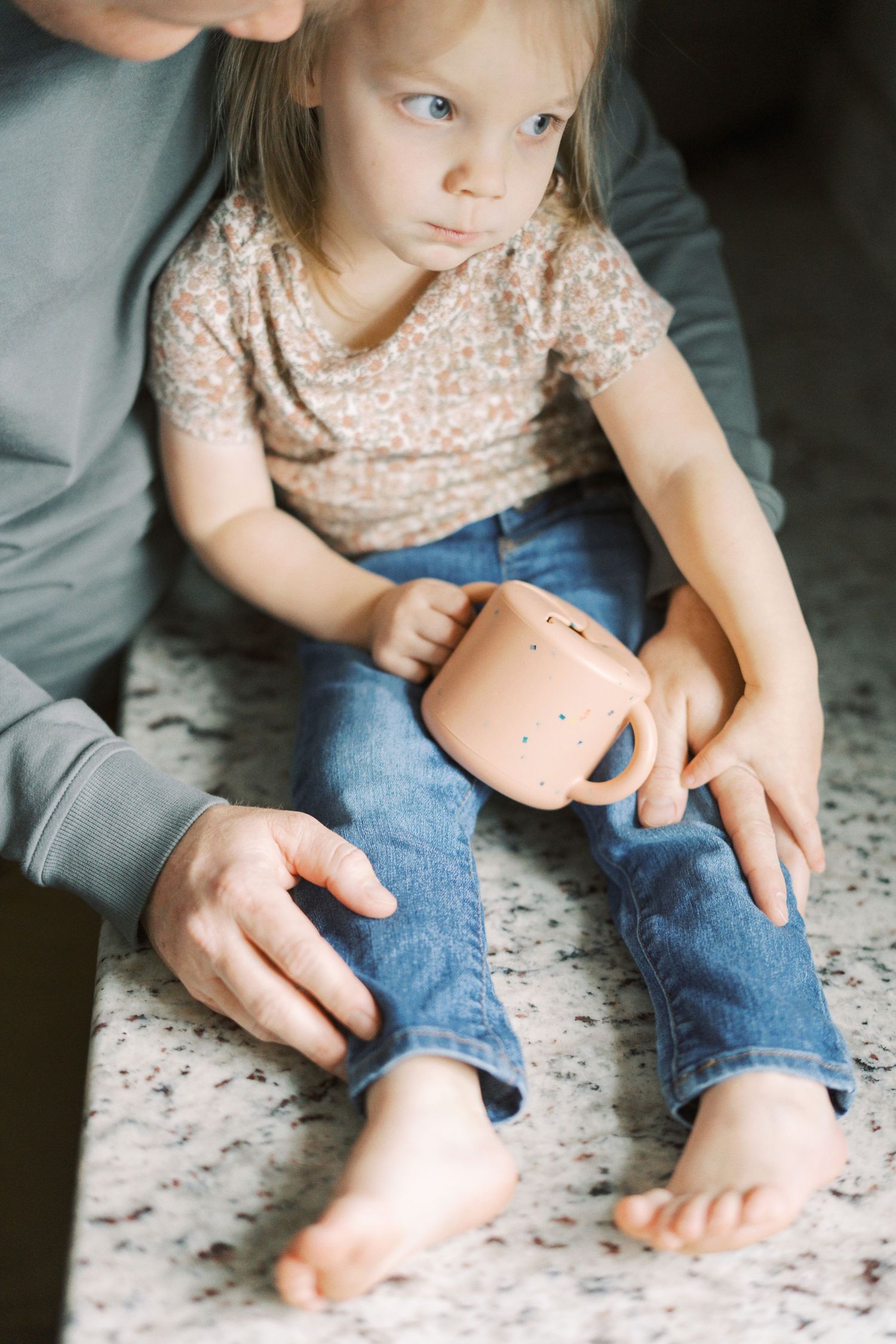 A close-up shot shows small bare feet and hands holding a pink cup while sitting on a textured carpet.