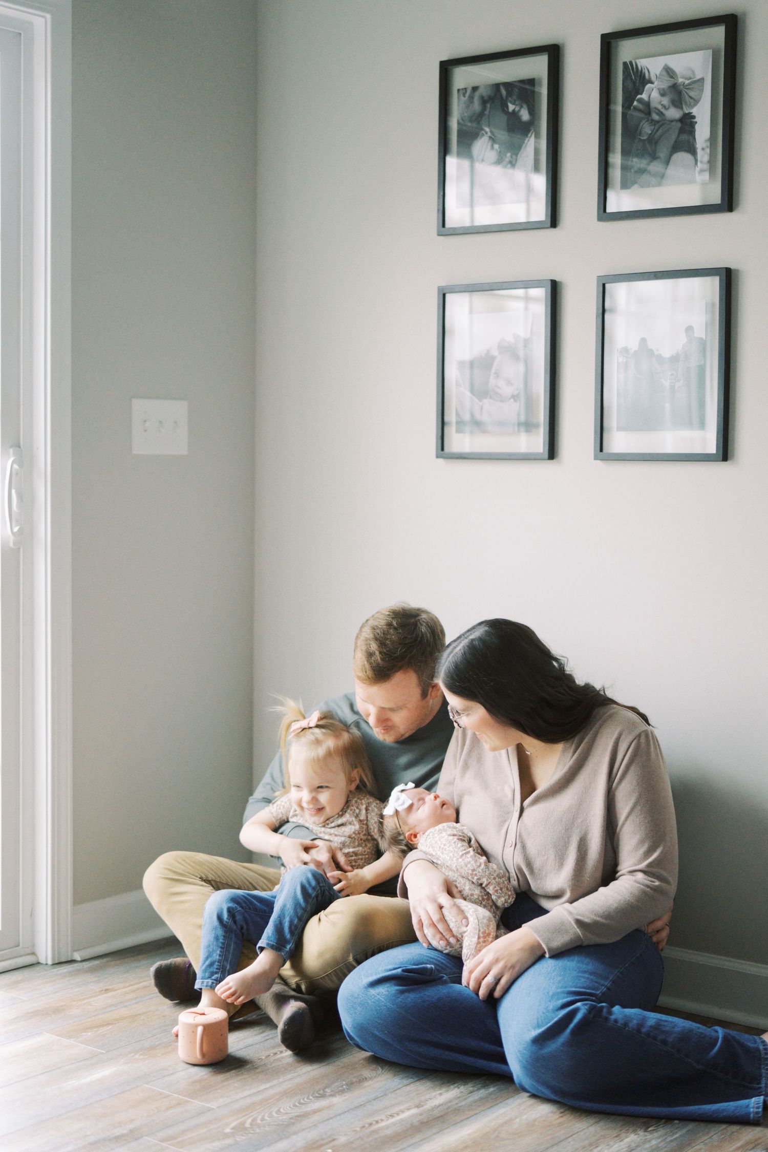A family sits together on hardwood flooring against a grey wall with framed photographs.