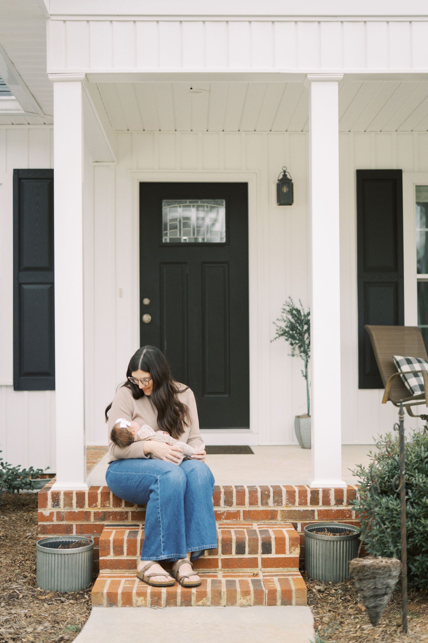 A person sits on brick porch steps of a white house with black doors and potted plants.