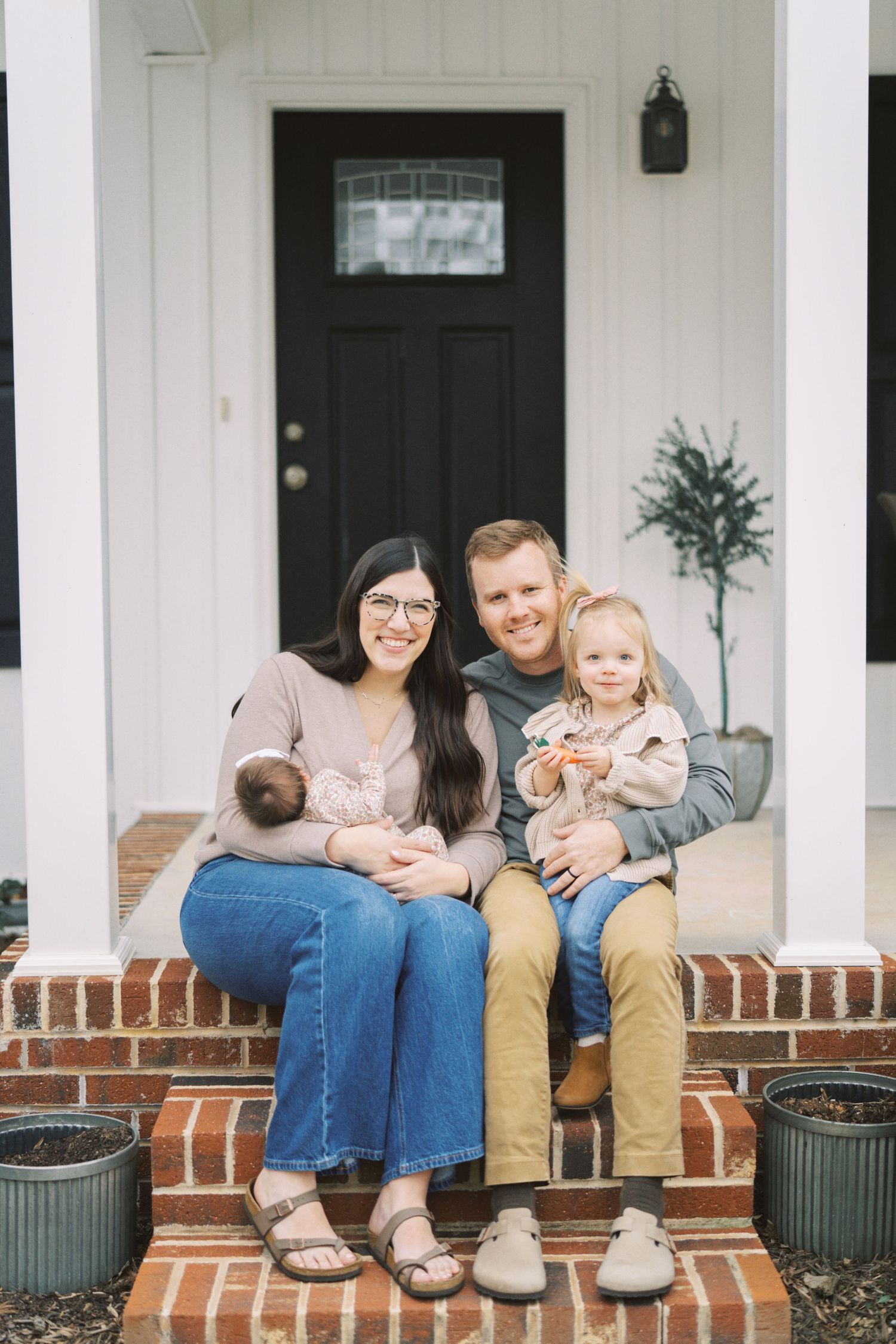 Family sitting together on brick front porch steps of white house with black door.