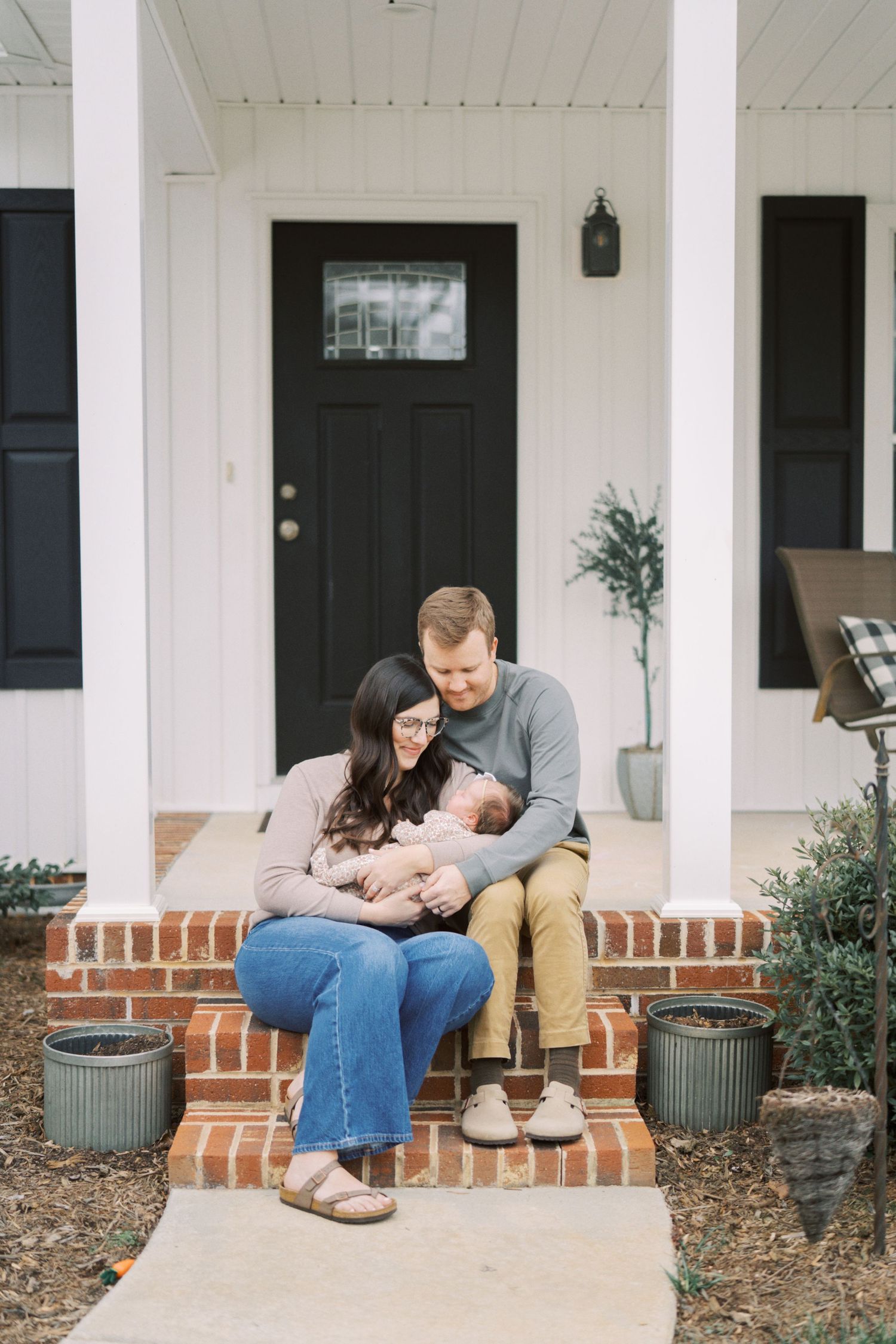 A couple sits together on brick porch steps of a white farmhouse with black shutters.