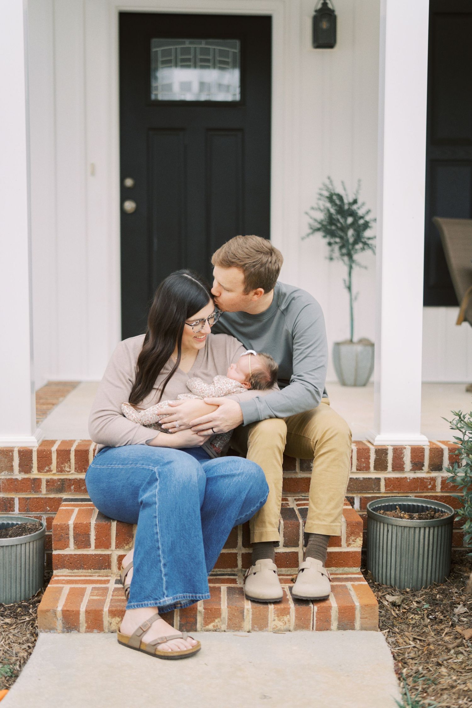 Family sits together with a newborn baby on their home's front brick steps near a black door and potted plants.