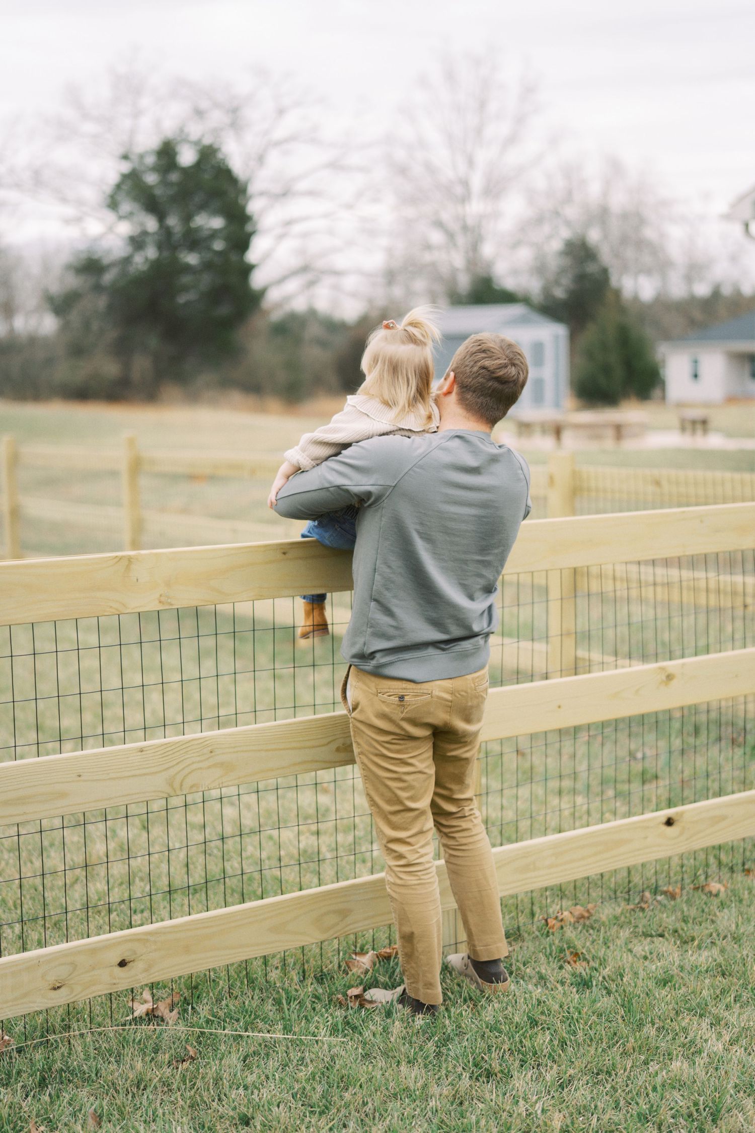 Two people stand at a wooden fence overlooking a rural pasture on an overcast day.