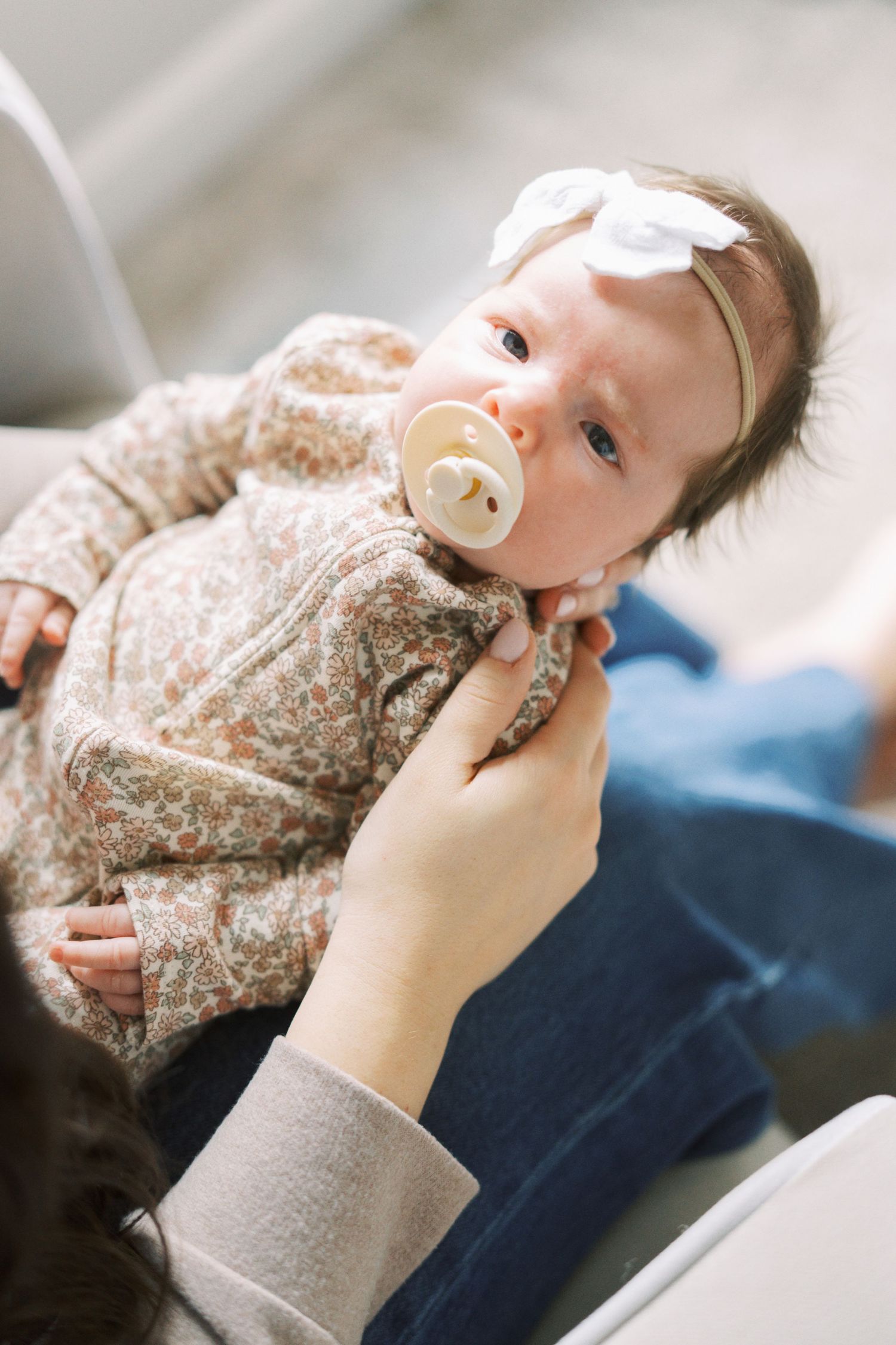 A baby wearing a floral print outfit with a pacifier is held by gentle hands.