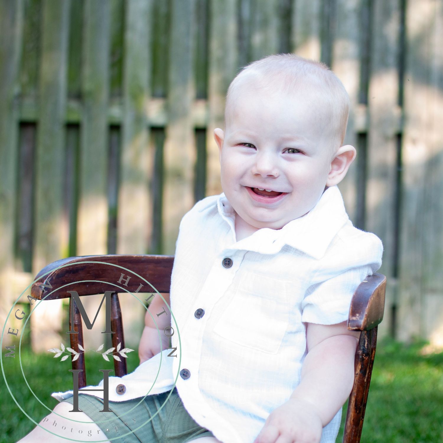A smiling one year old wearing a white collared outfit sits in a wooden chair in an outdoor setting for a portrait photograph.