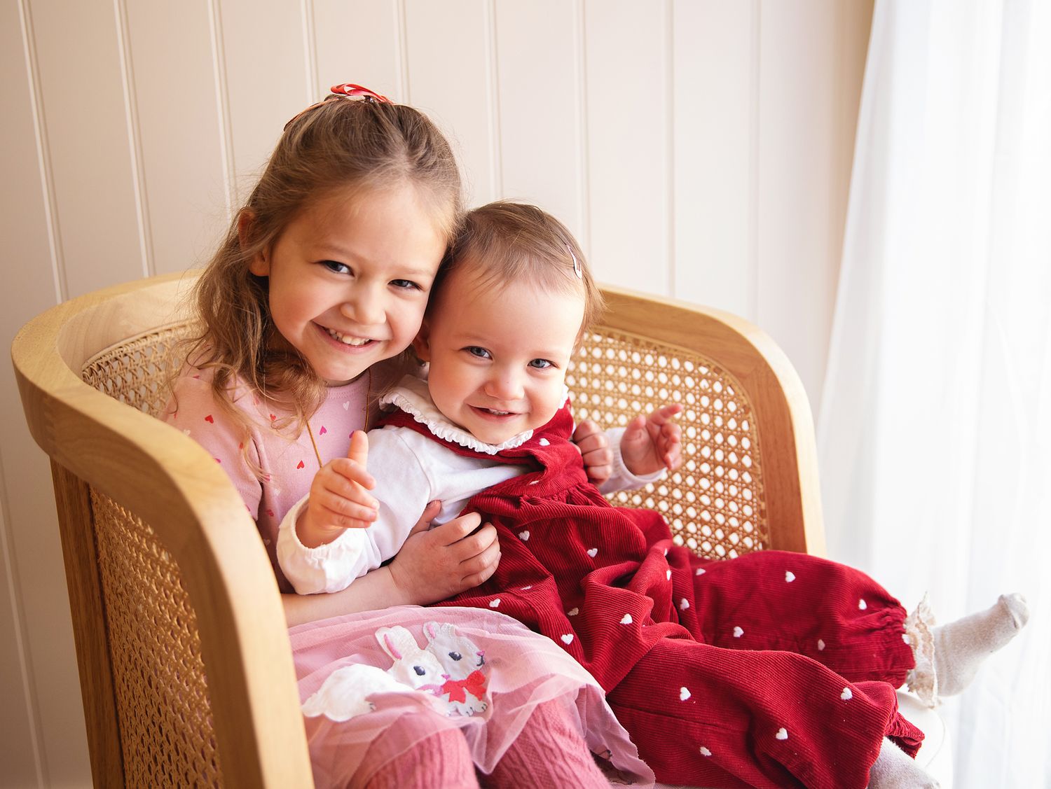 Two children sit together in a tan wicker chair, covered with a red blanket. During a baby photoshoot in Bexley