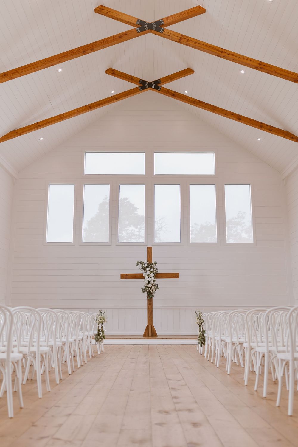 Modern white chapel interior with exposed wooden beams and white chairs arranged in rows facing a wooden cross altar.