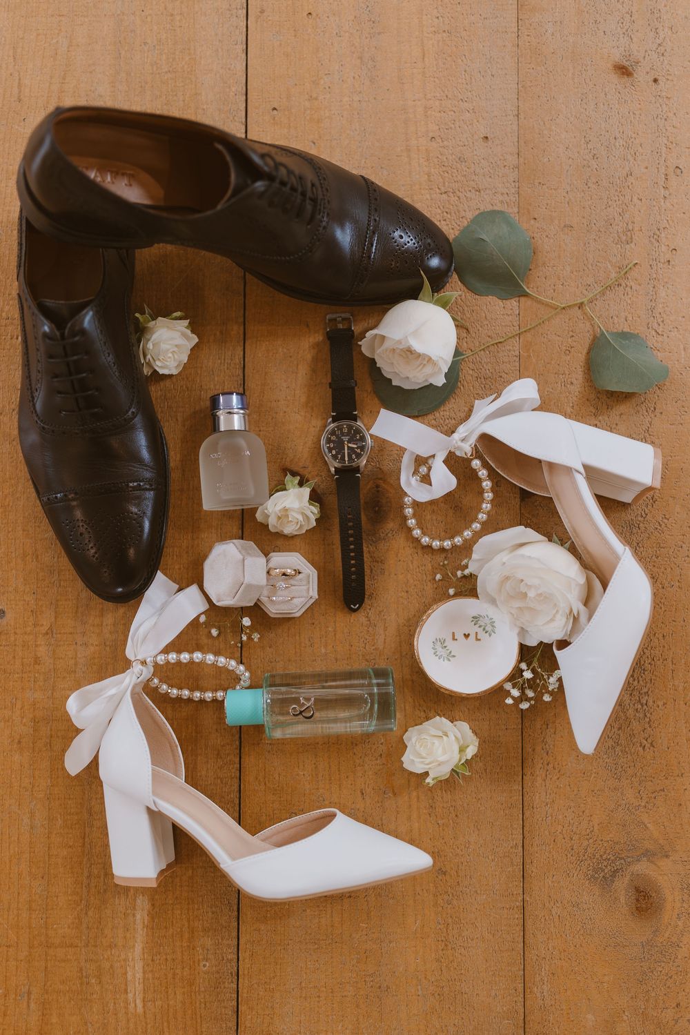 Wedding accessories including dress shoes, pearls, perfume bottles, and white roses arranged on wooden flooring.