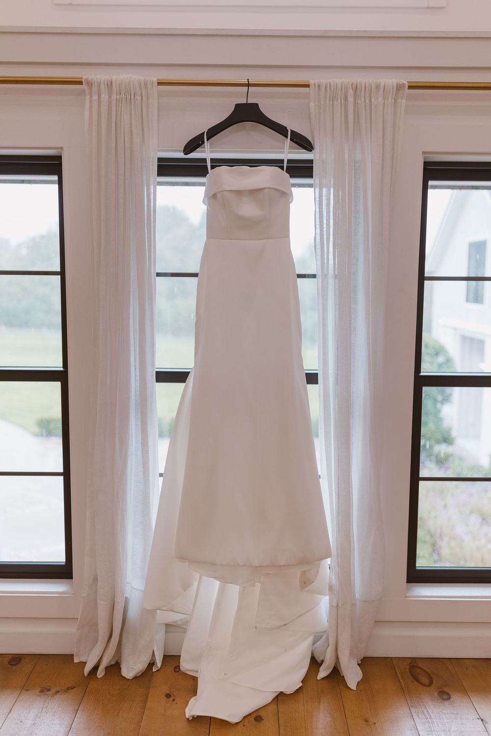 Elegant white wedding dress hanging on display between windows with sheer curtains in a sunlit room.