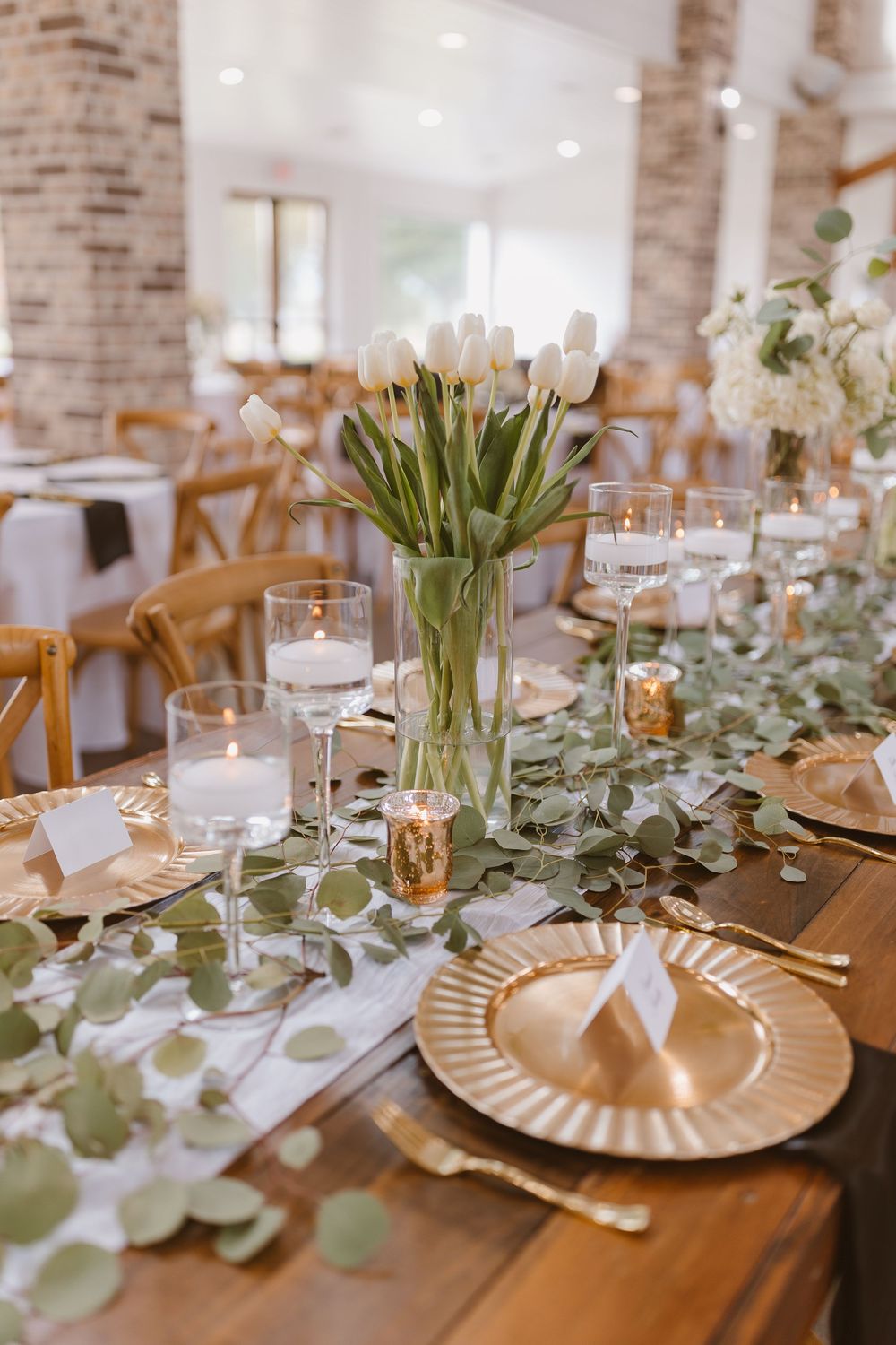Elegant wedding reception table setting with rustic brick walls, gold plates, and eucalyptus garland centerpieces.