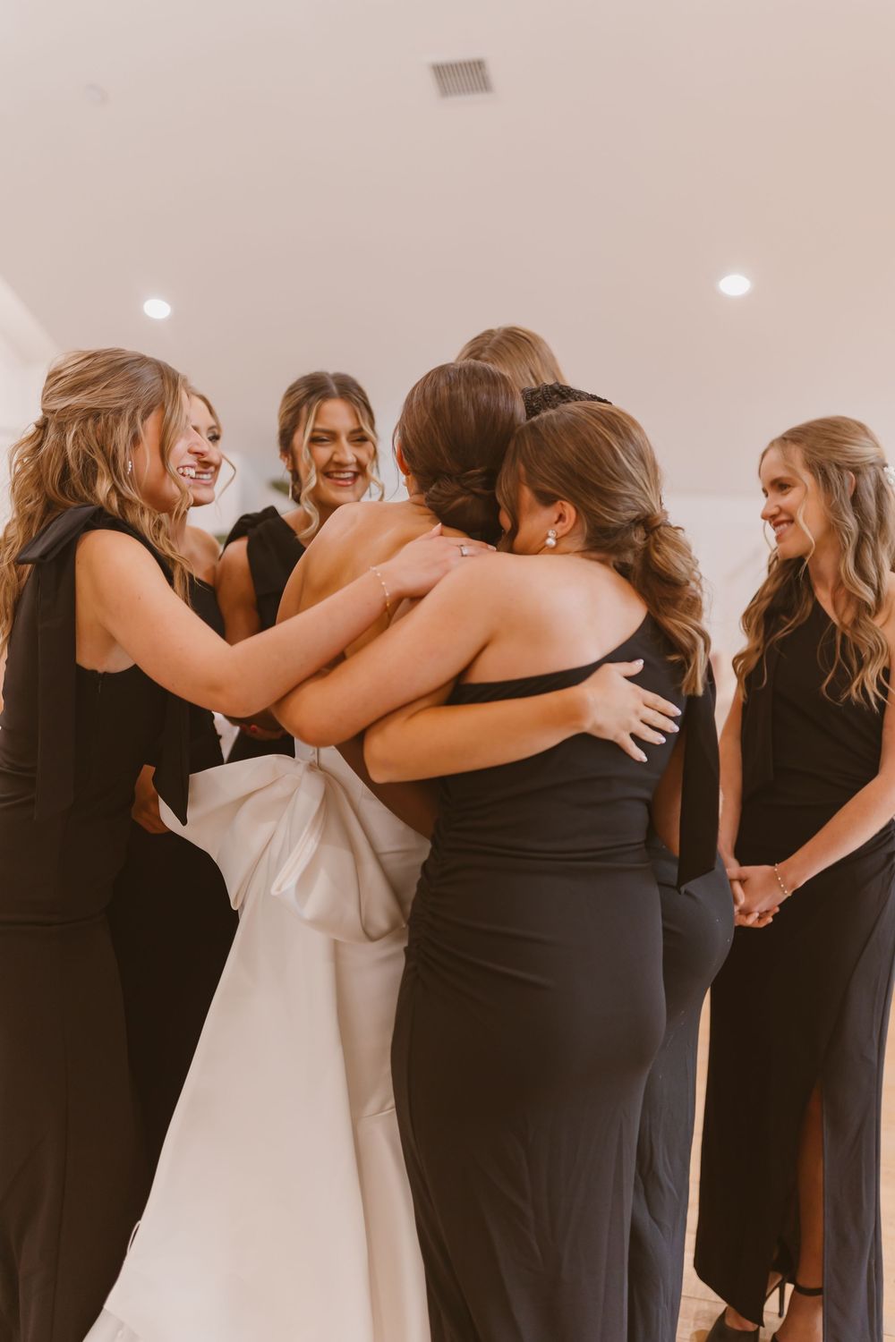 Bridesmaids in black dresses embrace the bride in a white gown during an emotional wedding moment.