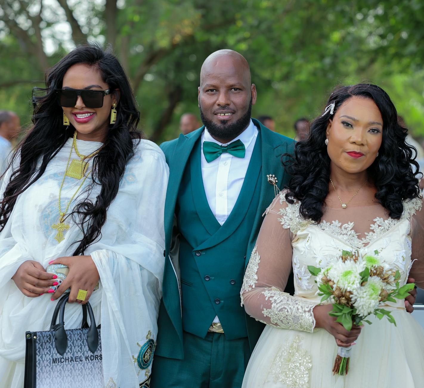 Elegant wedding photo showing three wedding guests in formal attire with teal suit and white dresses outdoors in garden setting.