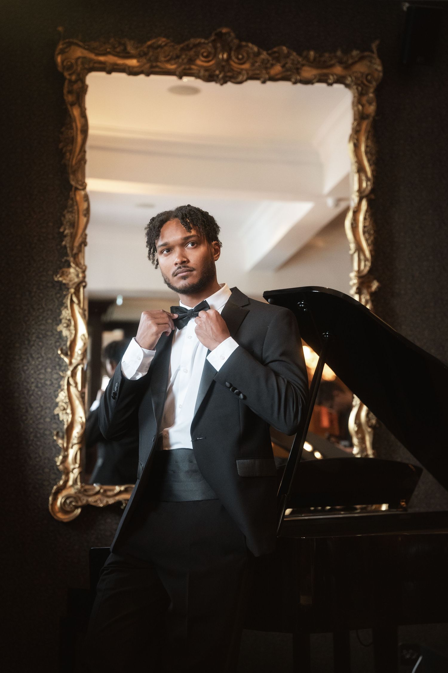 Stylish person wearing formal attire adjusts bowtie while standing in front of ornate gold mirror with grand piano visible.