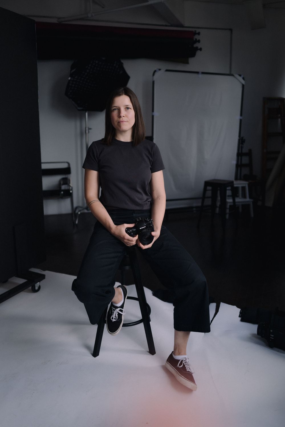 Professional photographer sits on stool in modern studio space wearing all black and holding camera in moody lighting setup.