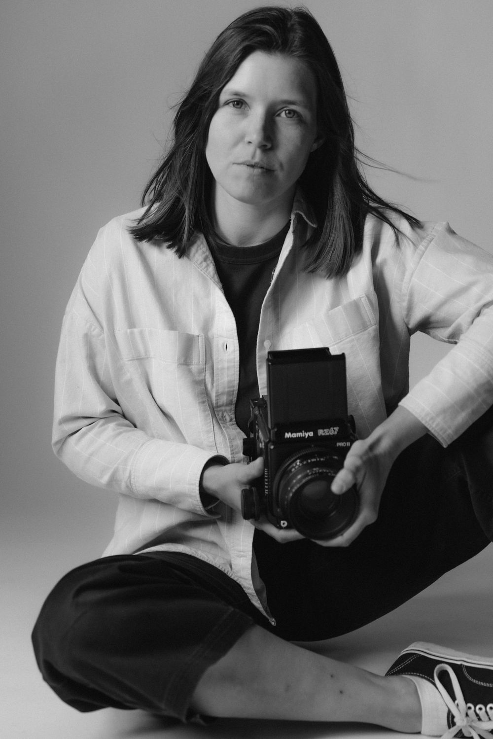 Black and white portrait of photographer in white jacket holding medium format camera while sitting cross-legged.