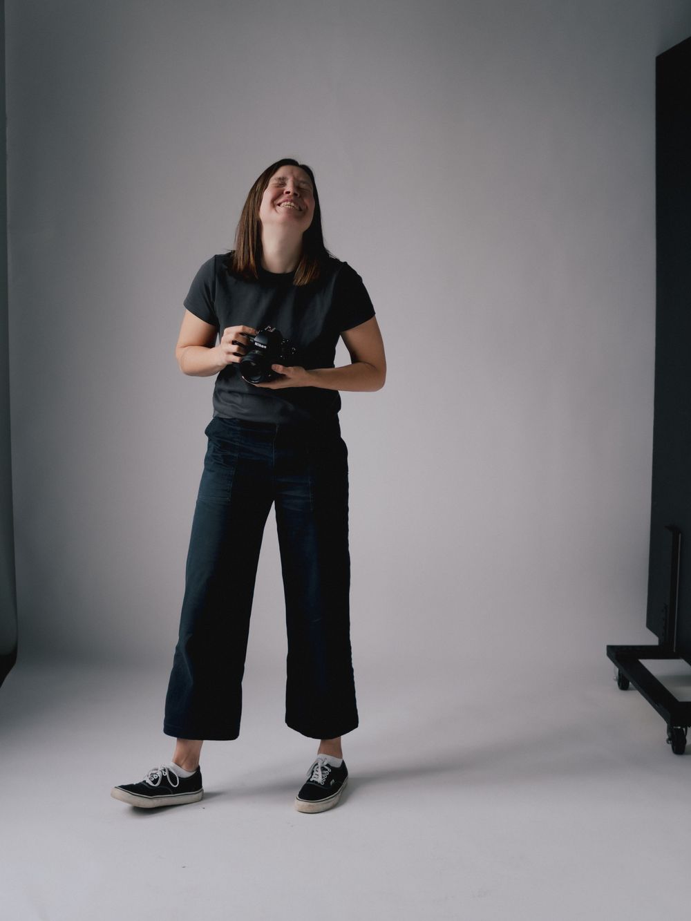A person in a casual black outfit holding a camera while standing in a minimalist studio setting with white background.