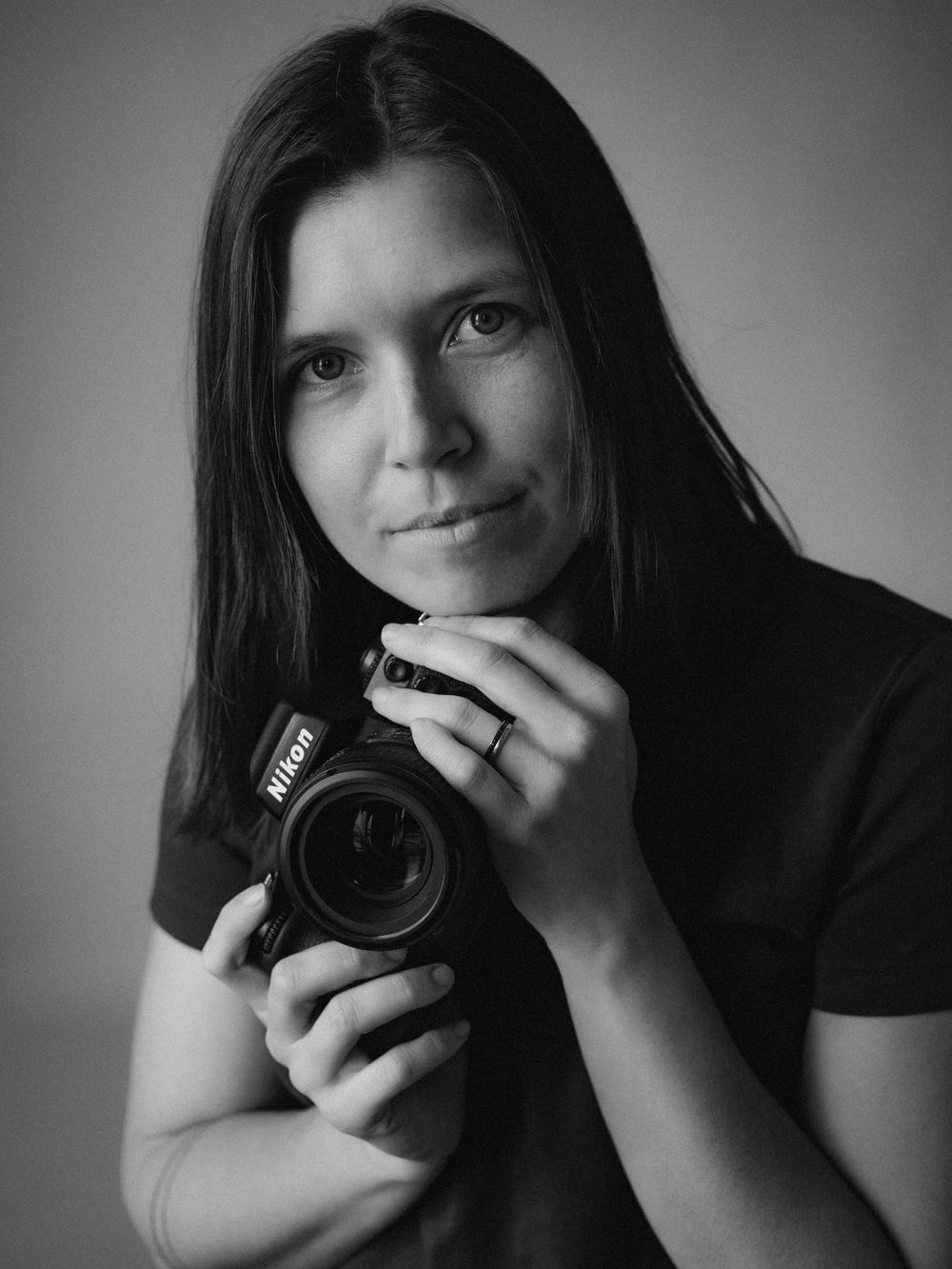 Black and white portrait of a photographer holding a Nikon camera while wearing dark clothing against a simple backdrop.