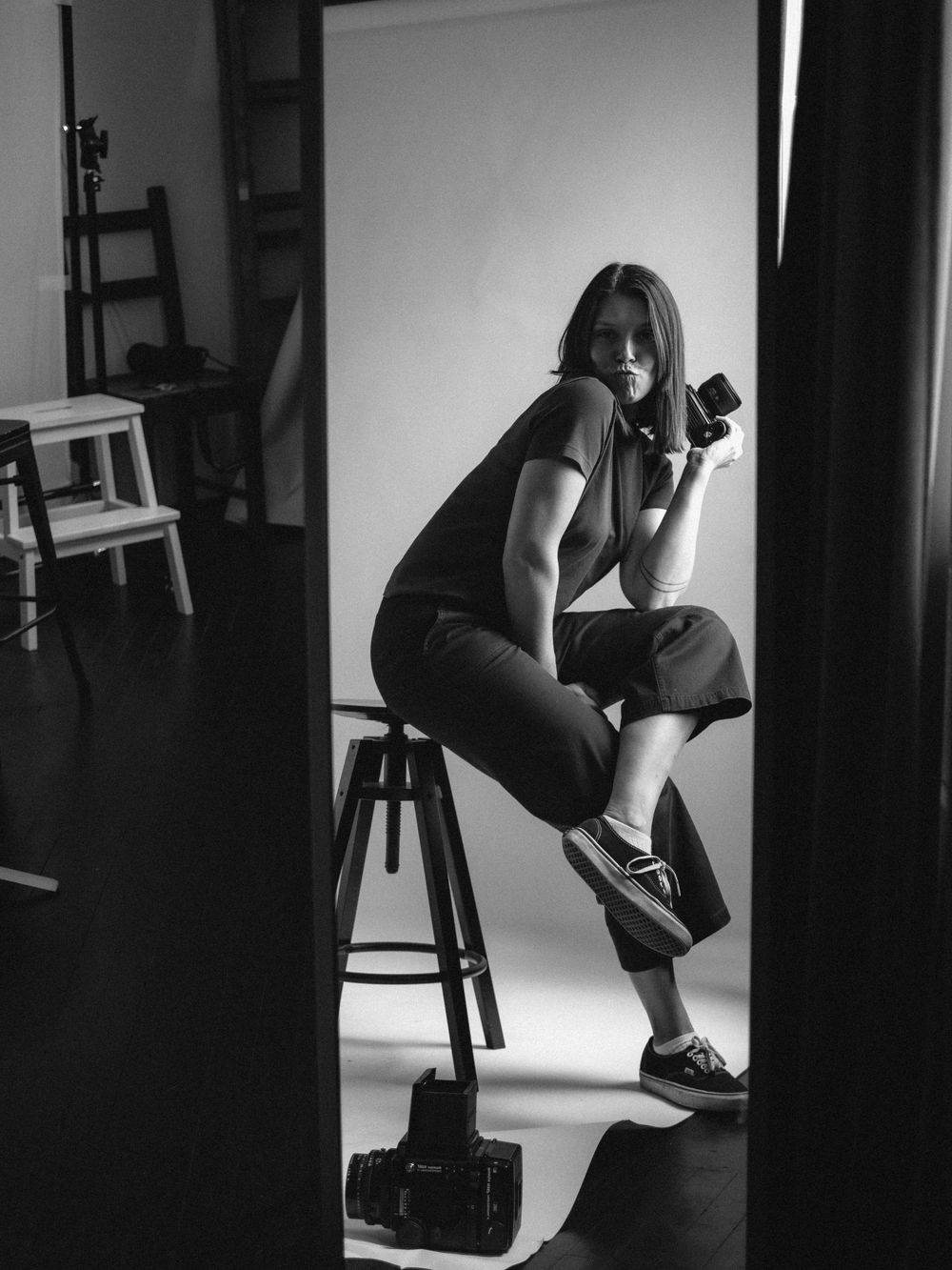 A black and white photograph of someone sitting on a stool taking pictures with a camera in a studio setting.