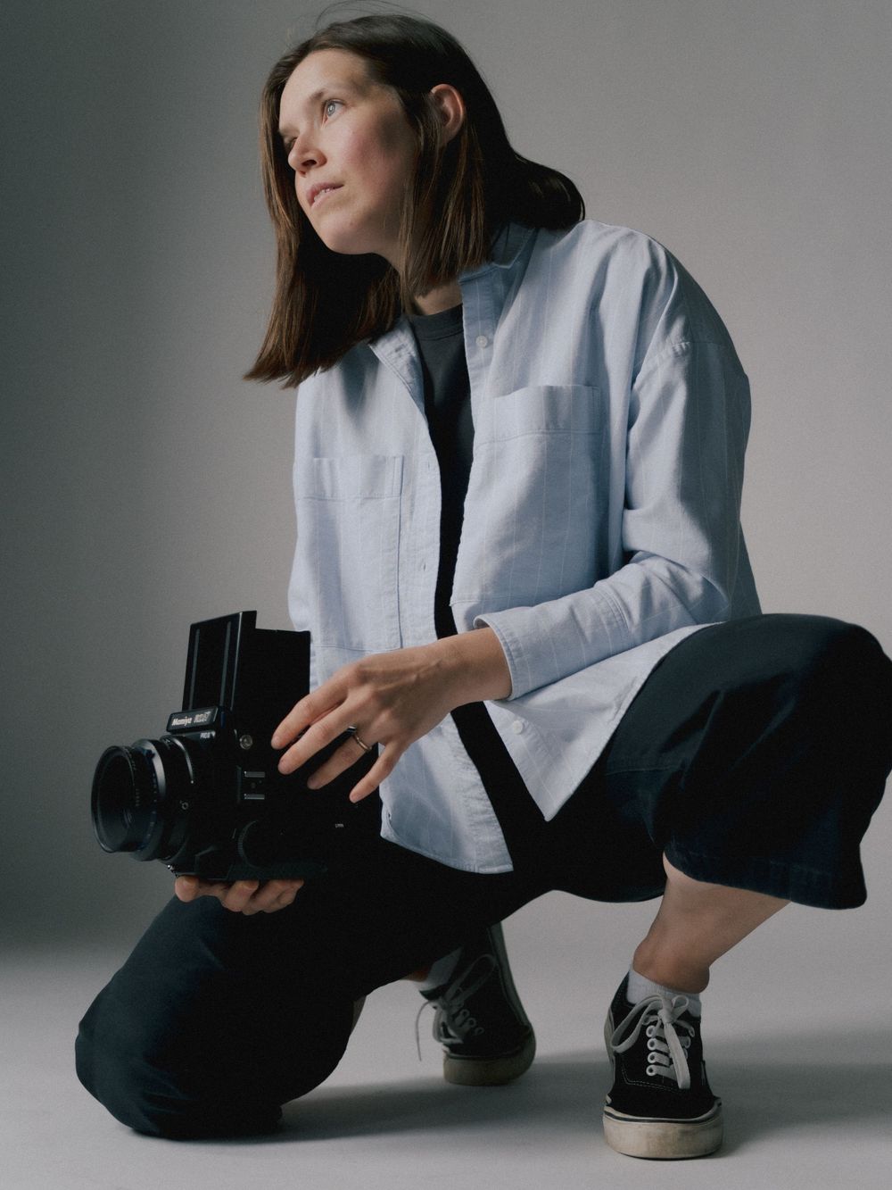 A photographer in white blazer and black pants sits casually on floor while holding a camera in studio setting.