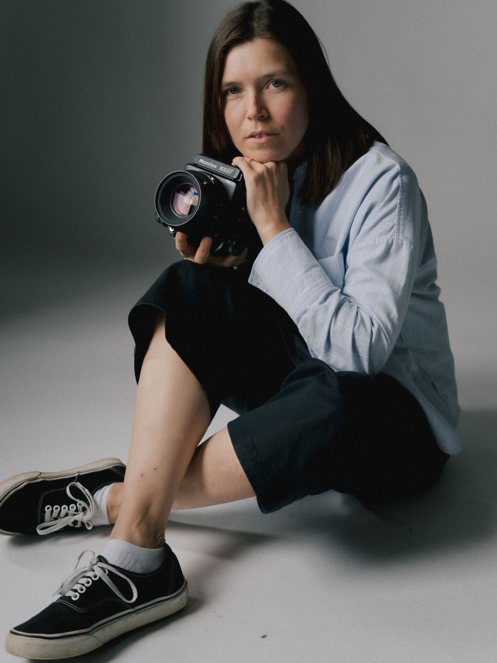 A photographer in casual attire sits on the floor with a DSLR camera while wearing black and white sneakers.