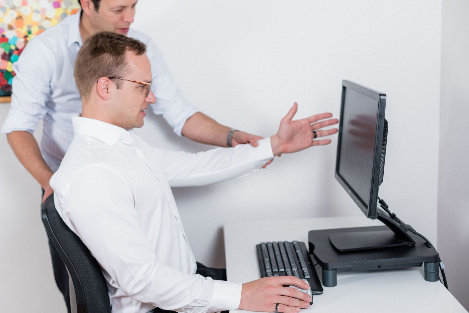 Professional office worker gestures at computer monitor while discussing work with colleague.