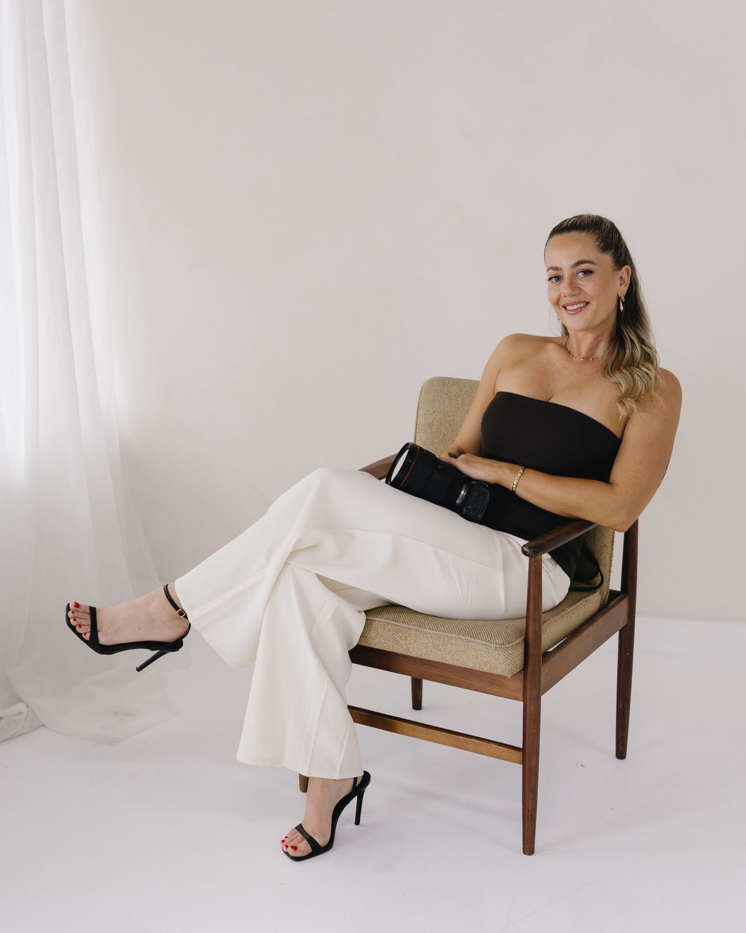 A person in a black strapless top and white pants sits relaxed in a mid-century modern armchair against a white background.