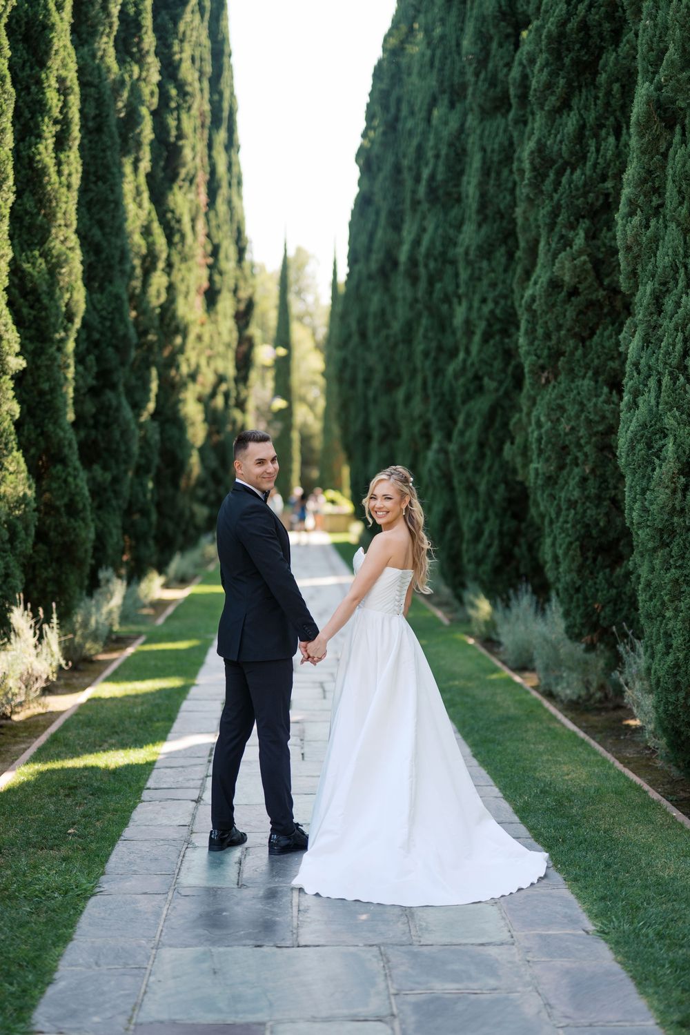 A wedding couple poses between tall cypress trees on a garden path lined with lush greenery and stone pavers.