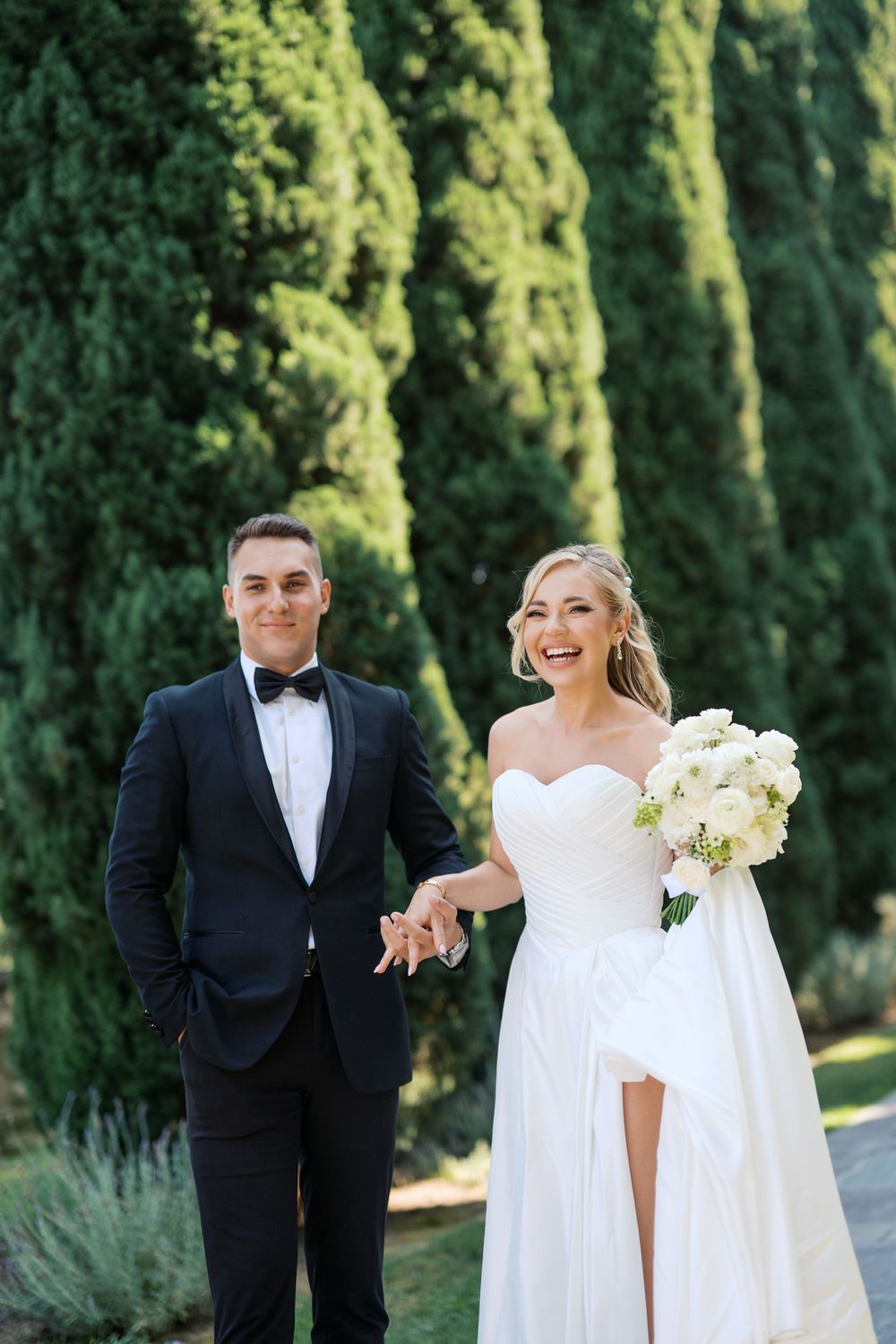 A wedding couple shares candid moments in a garden setting with tall cypress trees in the background.