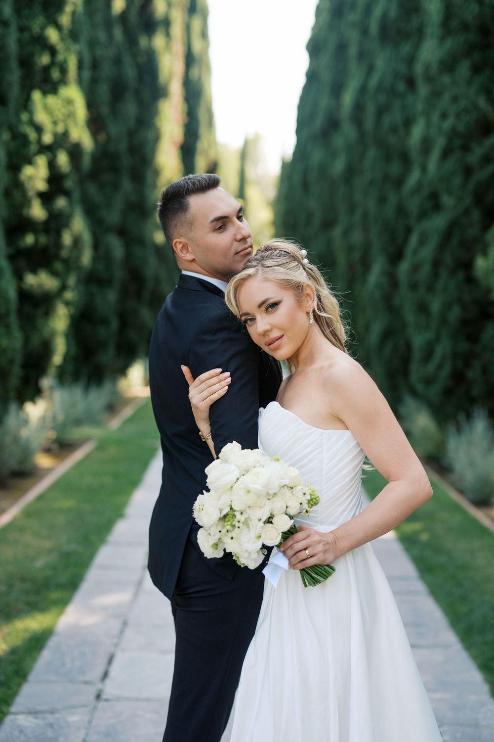 Romantic wedding couple poses along a cypress tree-lined path with white bouquet.