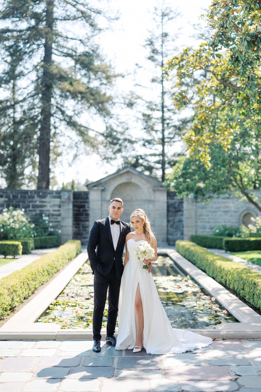 A couple poses in formal attire by a reflecting pool in a garden with tall evergreen trees.