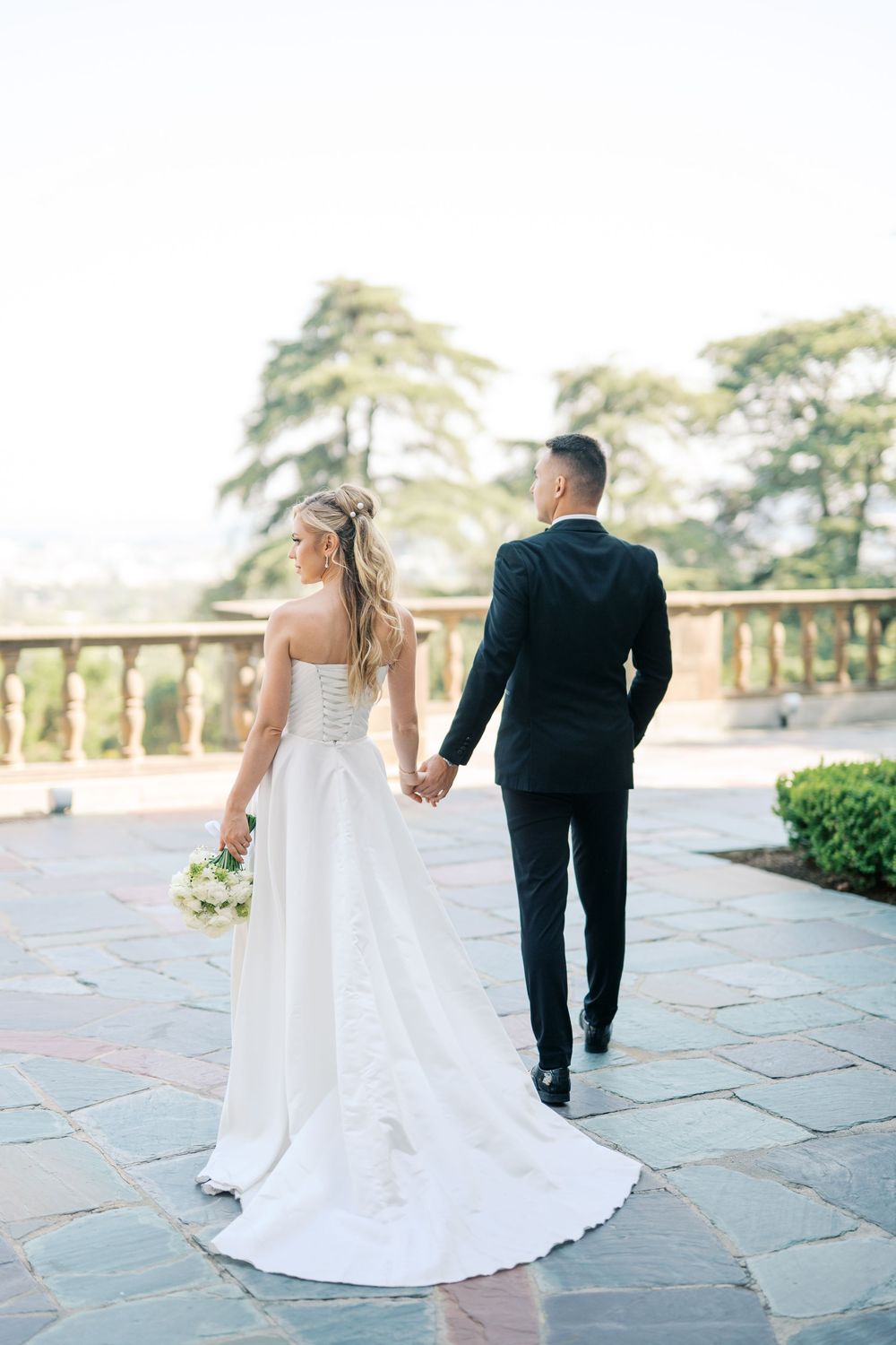 A couple holding hands while walking on a stone terrace with scenic mountain views in the background.