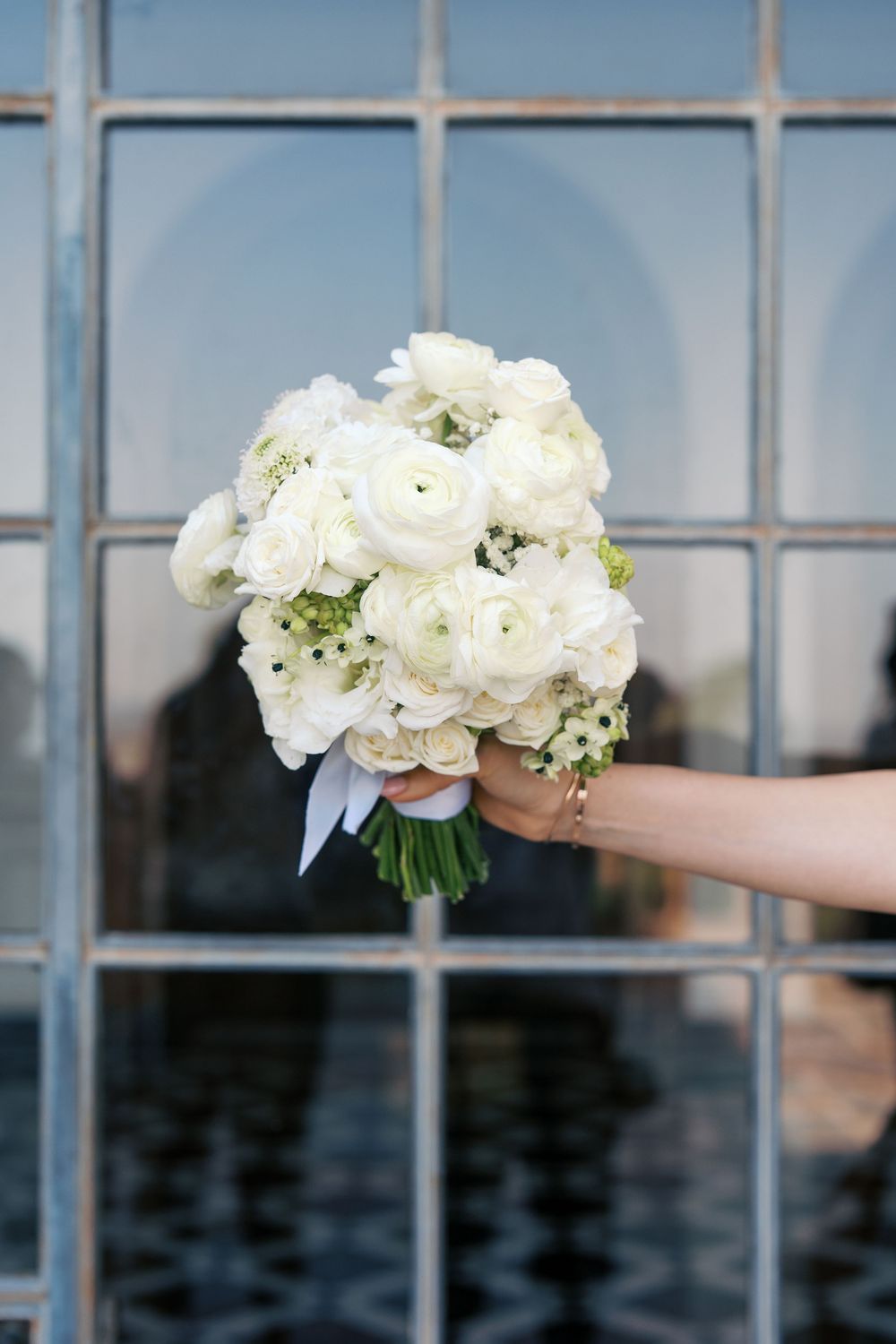 A delicate white bouquet of ranunculus and roses held against a window pane.