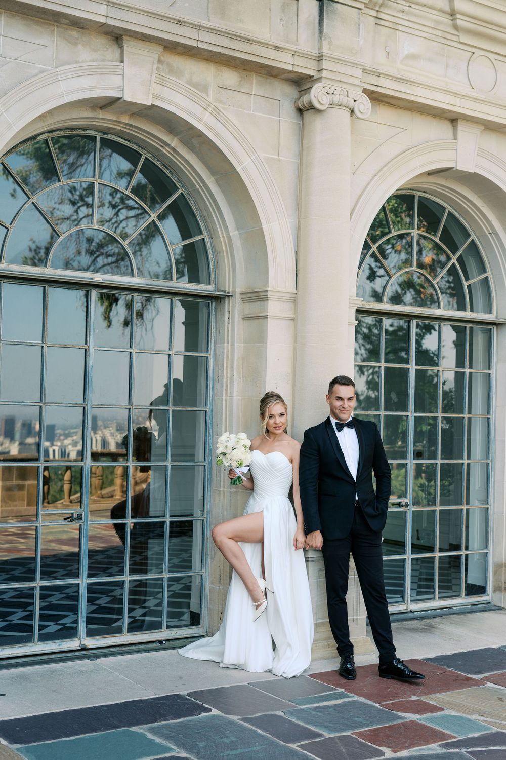 A couple poses in formal attire against large arched windows of an elegant stone building.