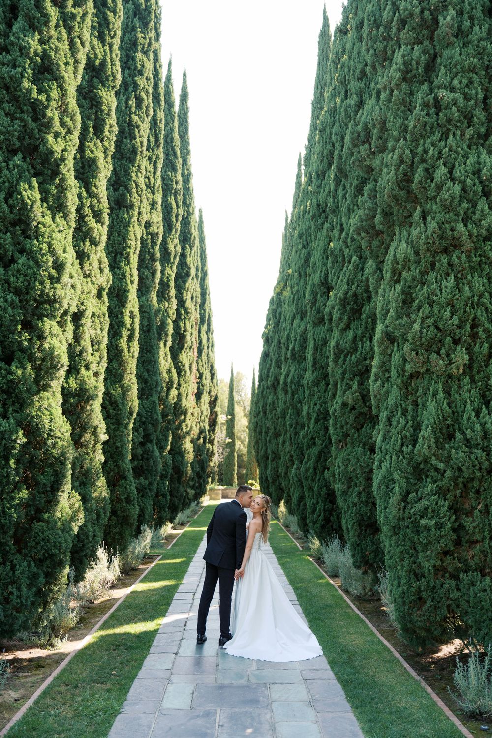 A dramatic walkway lined with tall cypress trees leads to a couple on their wedding day.