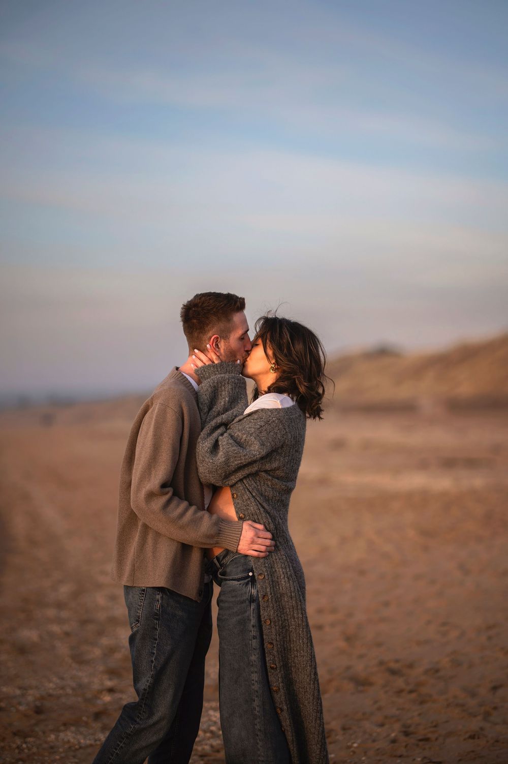 A romantic couple shares an intimate embrace while standing in a desert landscape at dusk.