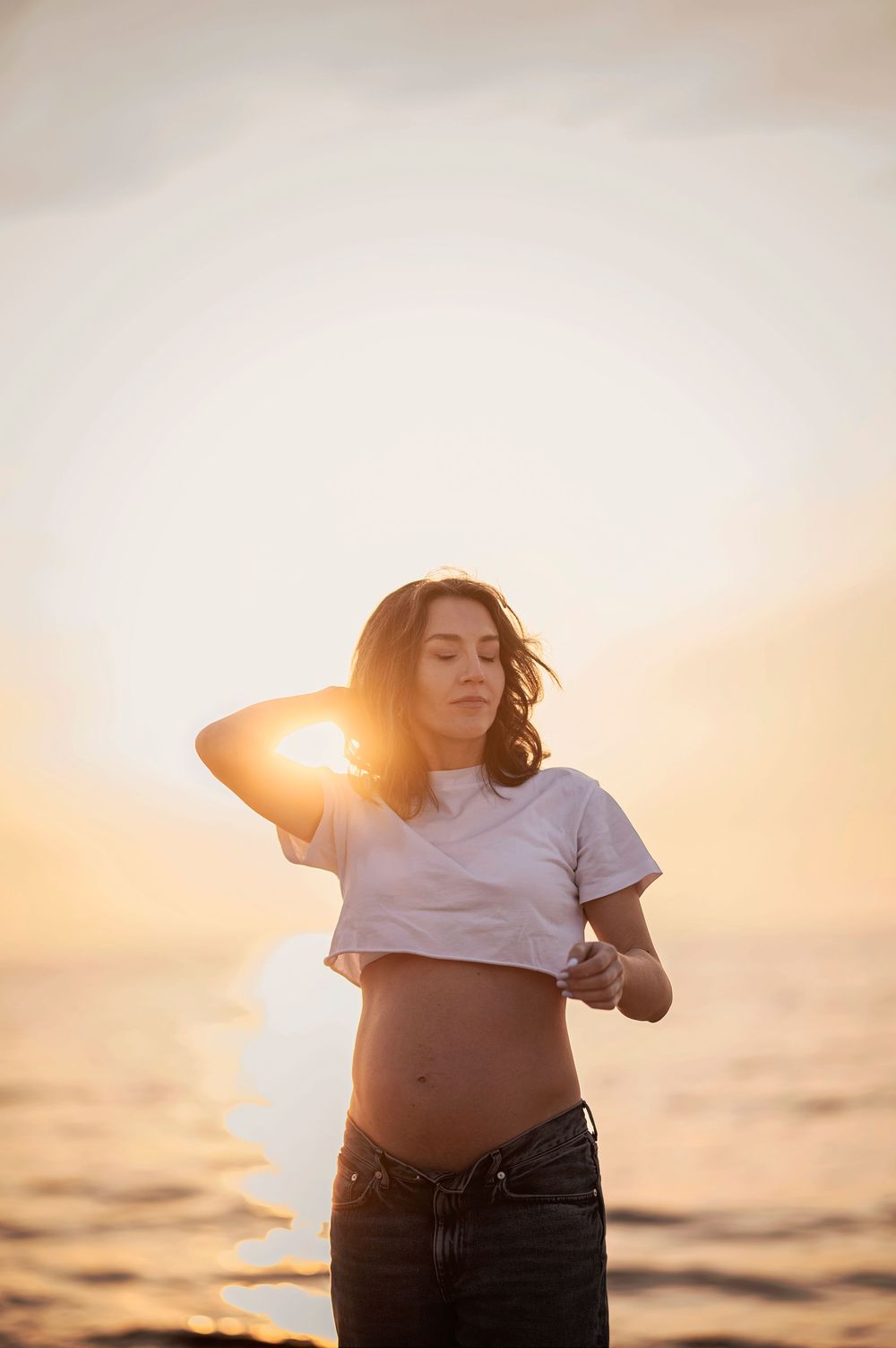 Silhouette against golden sunset at beach wearing cropped white t-shirt and jeans with wind-swept hair.