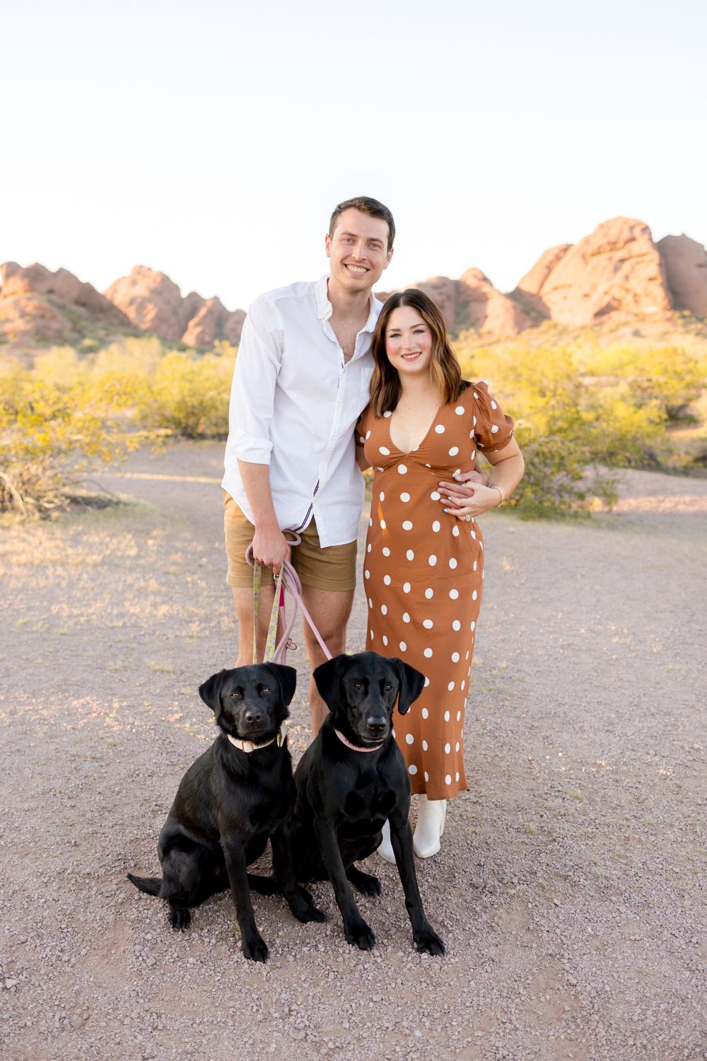 A couple poses outdoors in the desert with their two black Labrador dogs during golden hour.