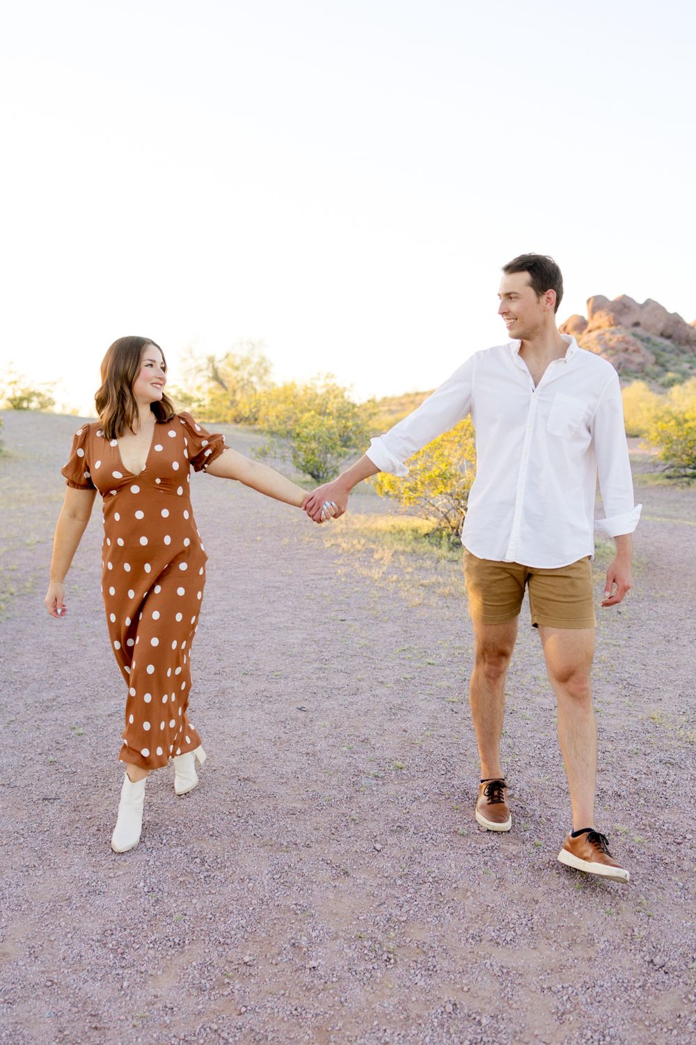 Couple holds hands while walking on desert path at sunset wearing casual outfits and white sneakers.