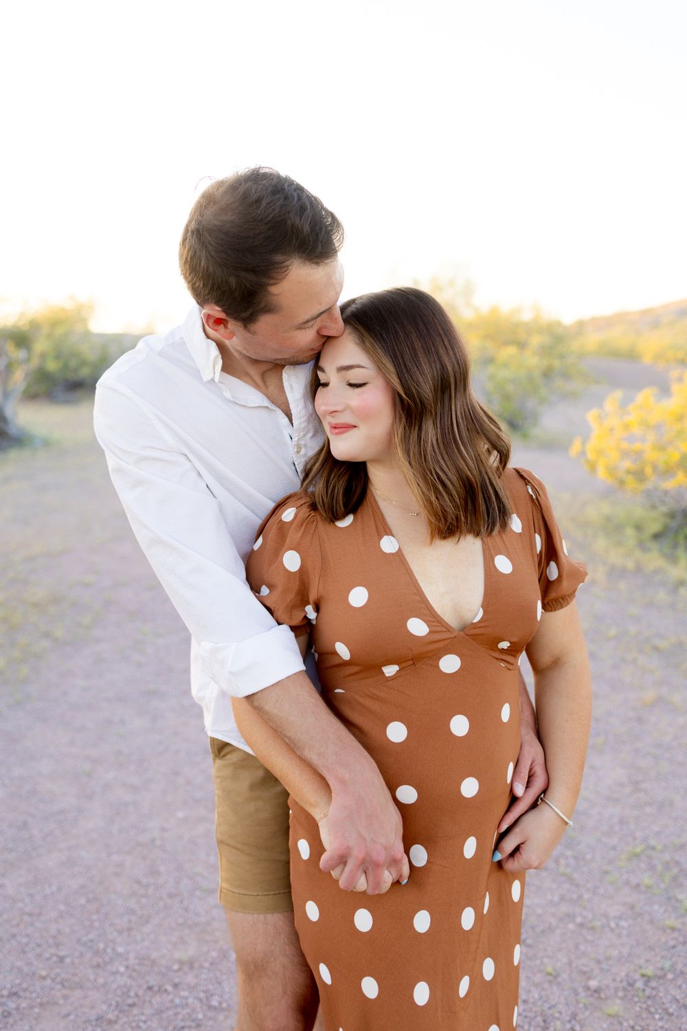 A couple embraces outdoors, with her wearing a brown polka dot dress and him in a white shirt and khaki shorts.