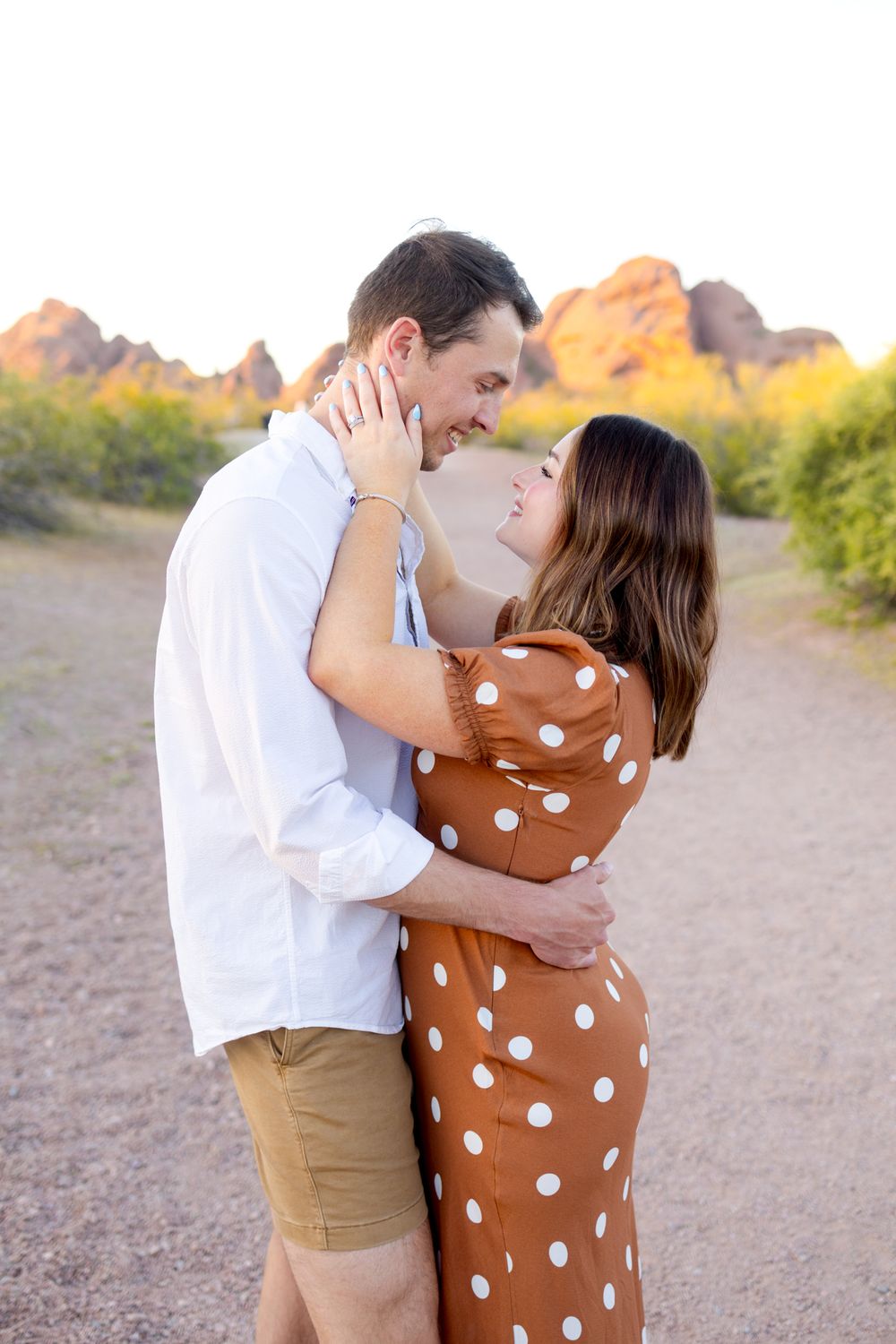 A romantic couple embraces in a desert landscape at sunset, with rocky mountains and desert plants in the background.