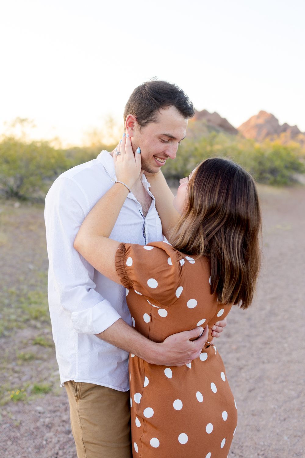A romantic couple embraces on a desert trail at sunset with mountains in the background.