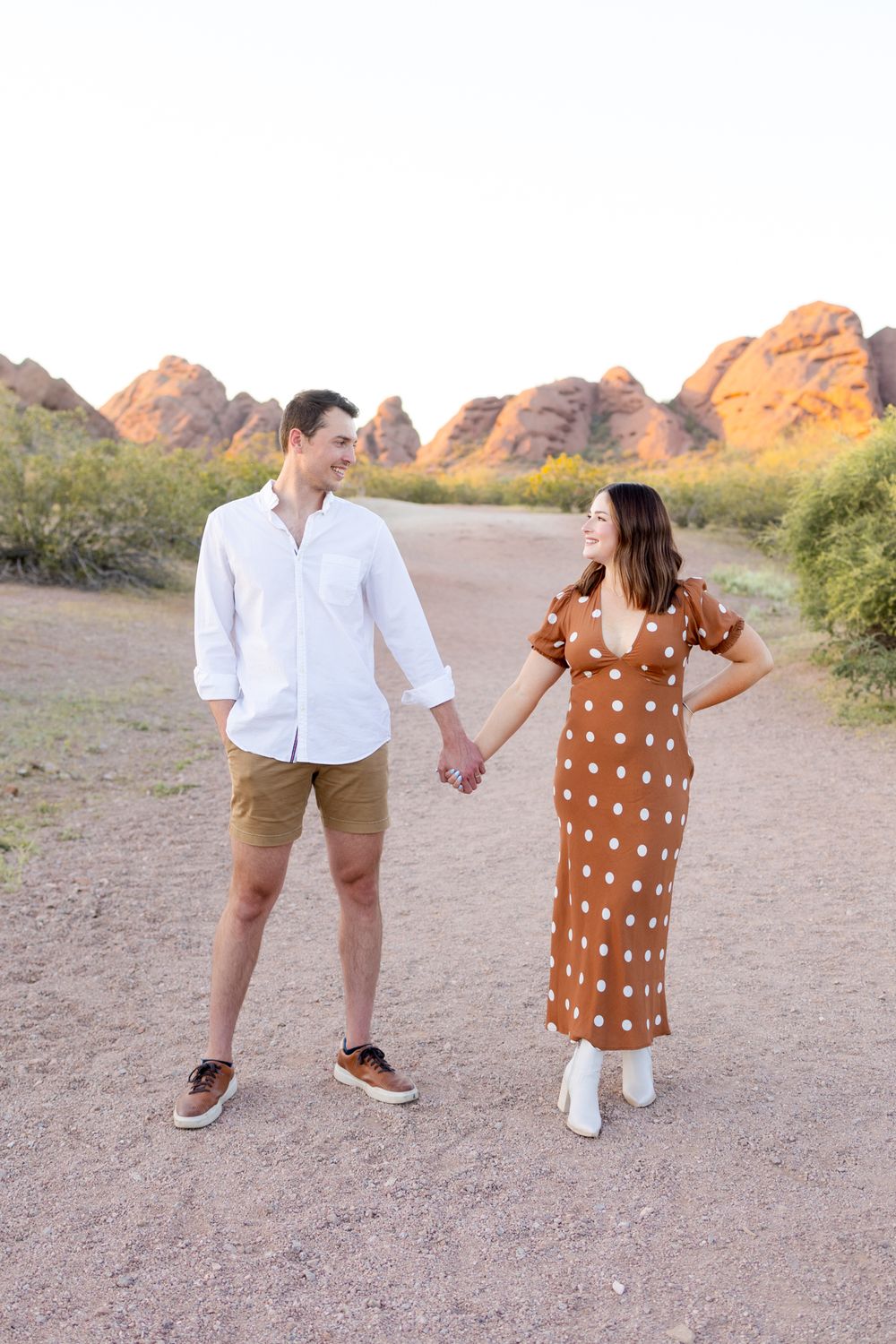 A couple holds hands while walking on a desert trail with red rock mountains glowing in the sunset.