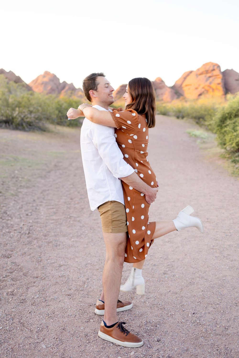 A romantic couple shares a playful embrace on a hiking trail with desert mountains in the background.