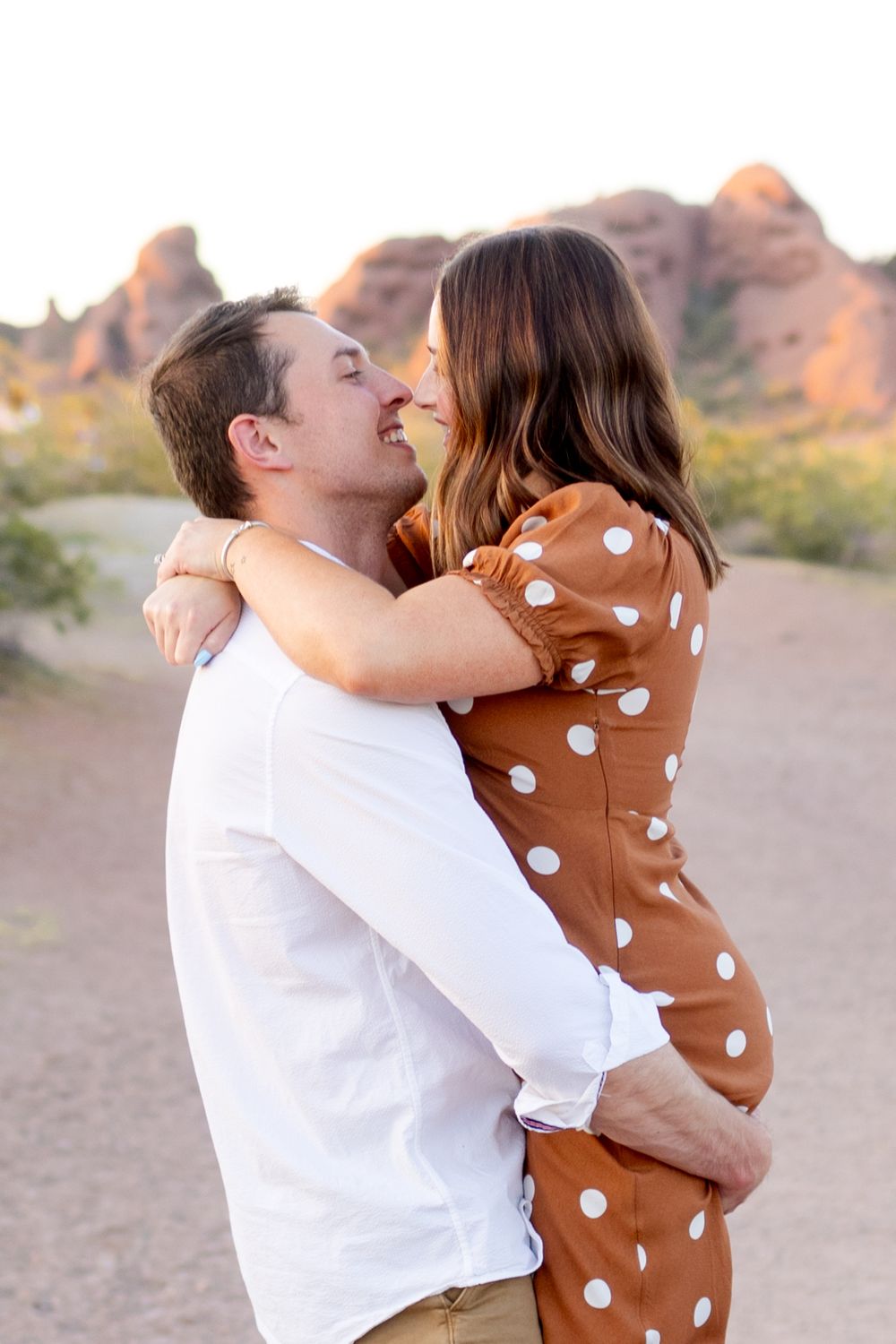 A couple embracing and kissing in a desert landscape with mountains in the background at sunset.