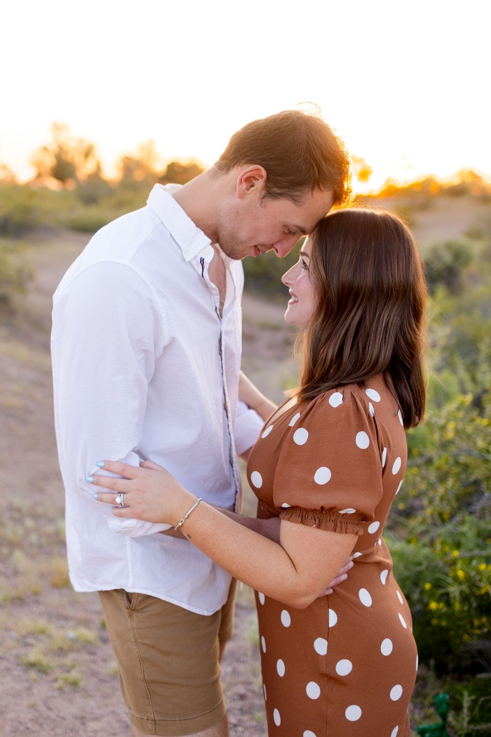 A couple shares an intimate moment at sunset in a desert landscape, her wearing a brown polka dot dress and him in white.