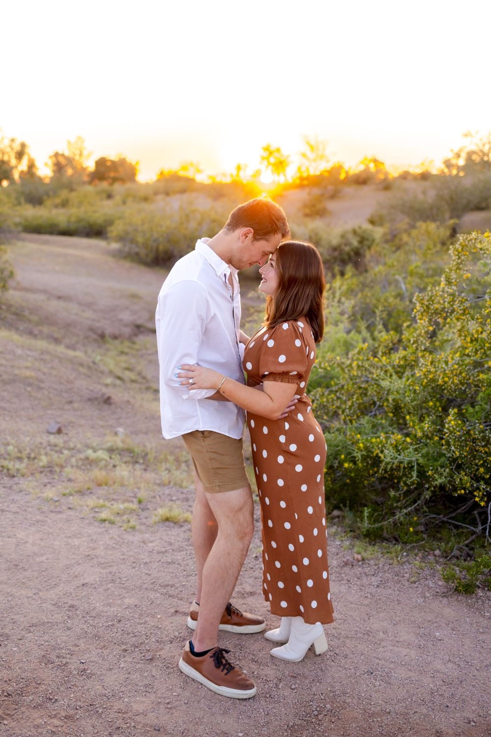 A couple embraces in a polka dot dress and casual wear during golden hour in a desert landscape with wildflowers.